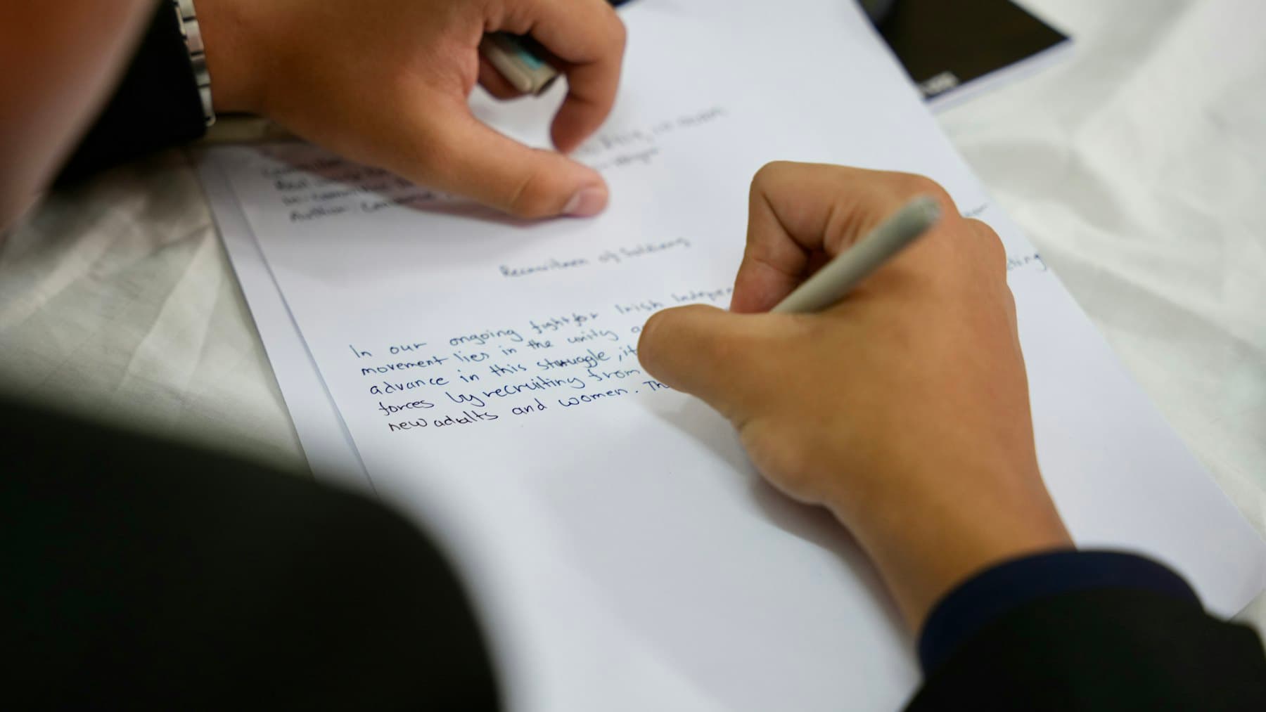 Two students working on a handwritten essay draft, one holding the page steady while the other writes.